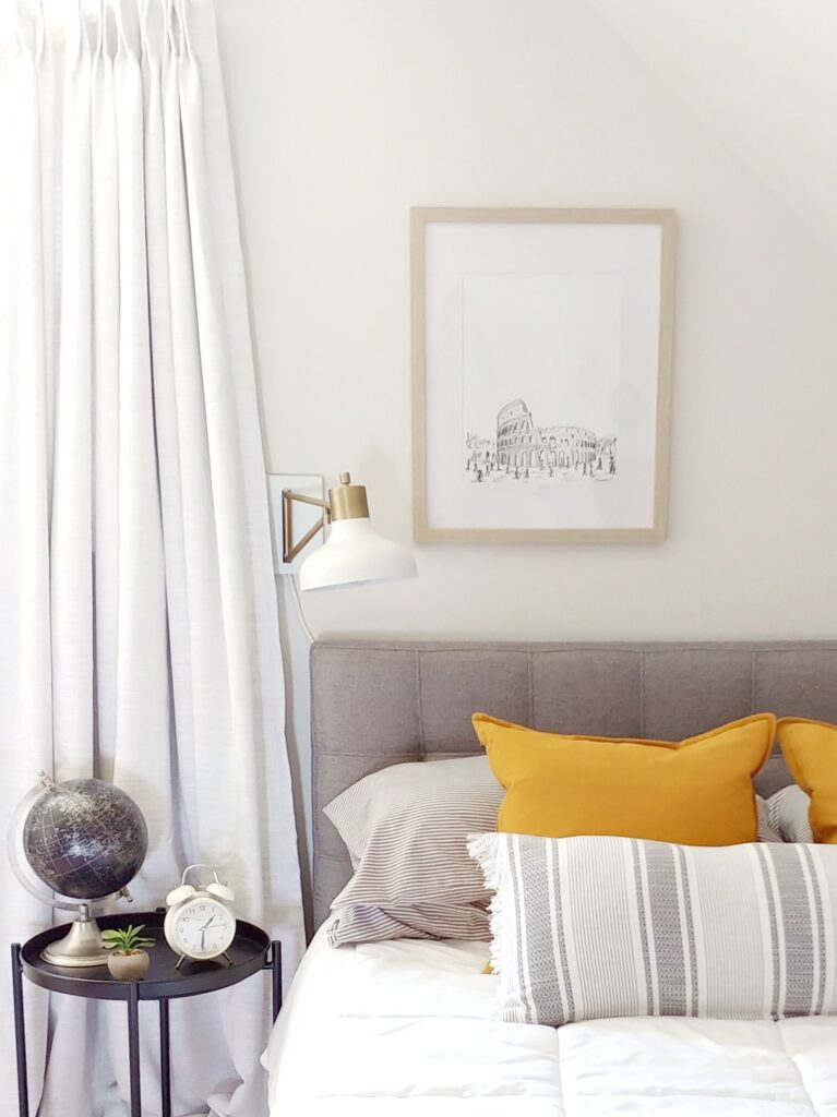 Grey Headboard with framed art of the Roman coliseum and a white scone on the wall and a black globe on the side table.