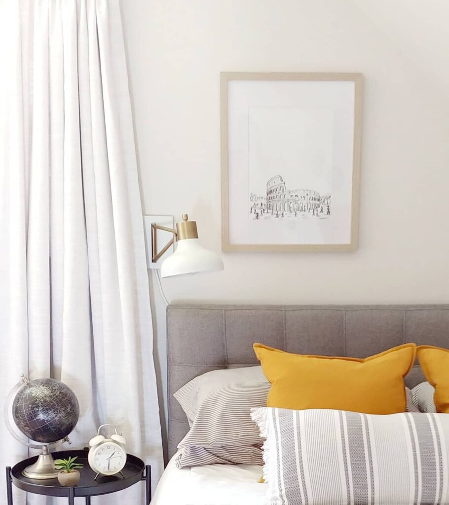 Grey headboard with striped lumbar pillow and a white scone on the wall over a black globe resting on the side table.