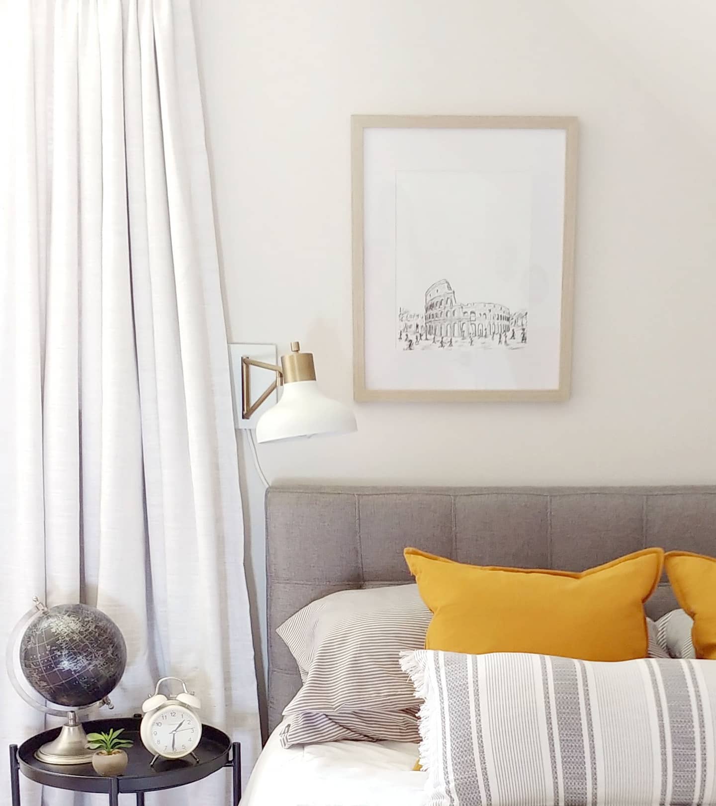 grey headboard with a framed drawing of the roman coliseum with a white scone on the wall and a black globe on the side table